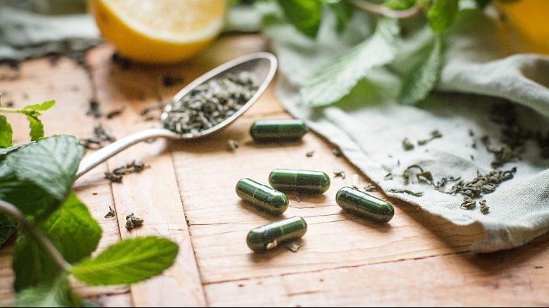 tigovit Grüntee-Extrakt Kapseln mit EGCG auf Holztisch neben Löffel mit losem Tee und frischen Minzblättern.