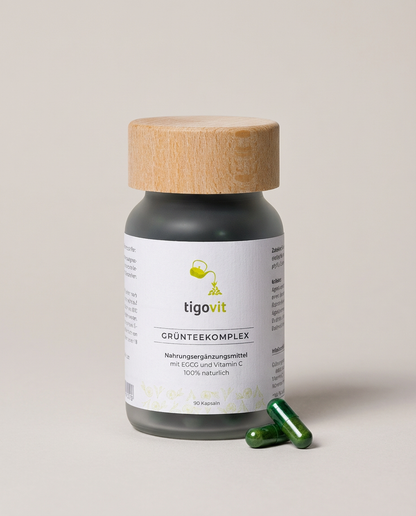 tigovit Grüntee-Extrakt Komplex Flasche (dunkles Glas, Holzdeckel) und zwei grüne Kapseln auf hellem Untergrund.